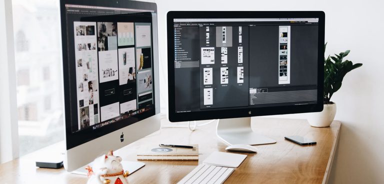 Two computer monitors on a wooden desk display various designs. One monitor shows different interface templates, while the other displays a detailed design workflow. A keyboard, a mouse, a notebook, and a potted plant are also on the desk. A window is in the background.