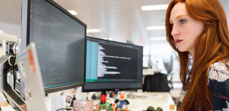 A person with long red hair is focused on a computer screen displaying code. They are in a modern office with additional monitors and various items on the desk, including small figurines. The background shows a bright, open workspace.