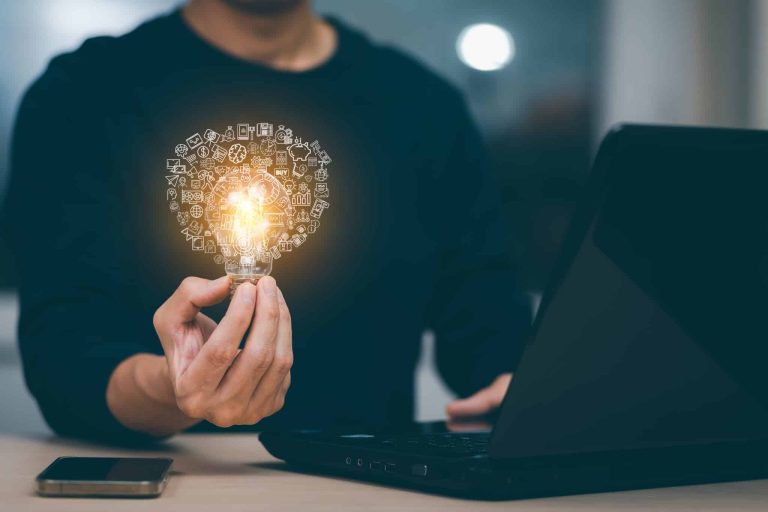 A person holding a glowing light bulb in their left hand while using a laptop. The light bulb is surrounded by various small icons, symbolizing ideas and technology. A smartphone lies on the table beside the laptop.