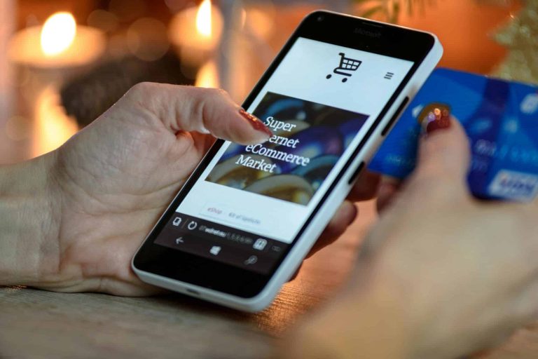 A person is holding a smartphone displaying an e-commerce website and a credit card in the other hand. The phone screen shows a shopping cart icon and text. The background is softly blurred with warm lighting.