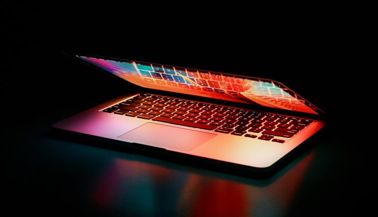 A partially open laptop with its screen glowing in vibrant colors of red, pink, and blue. The keyboard is illuminated by the light from the screen, creating a neon effect against the dark, black background. The laptop is positioned on a reflective surface.